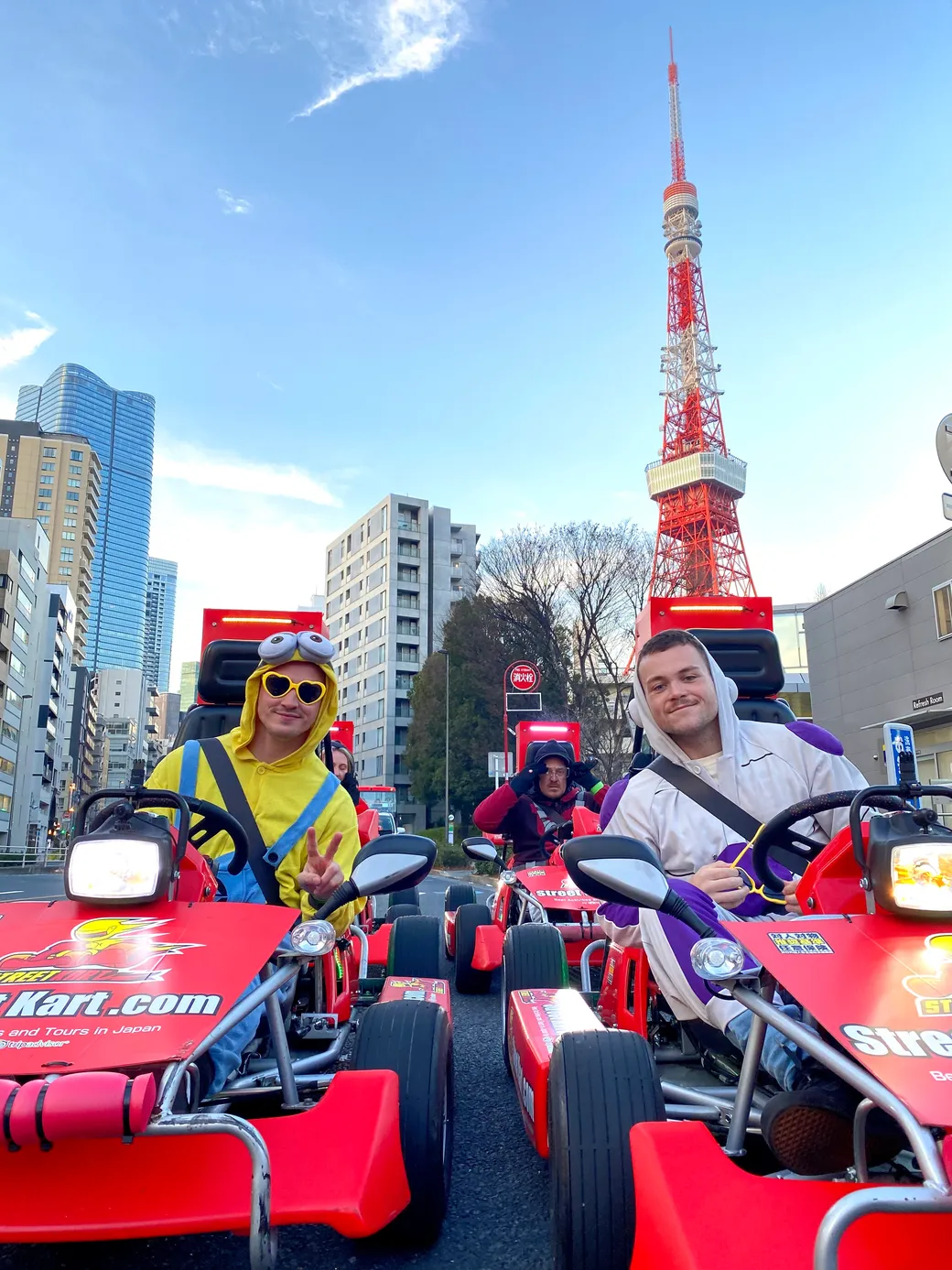 Three people in colorful costumes riding red go-karts on a city street, with Tokyo Tower visible in the background.