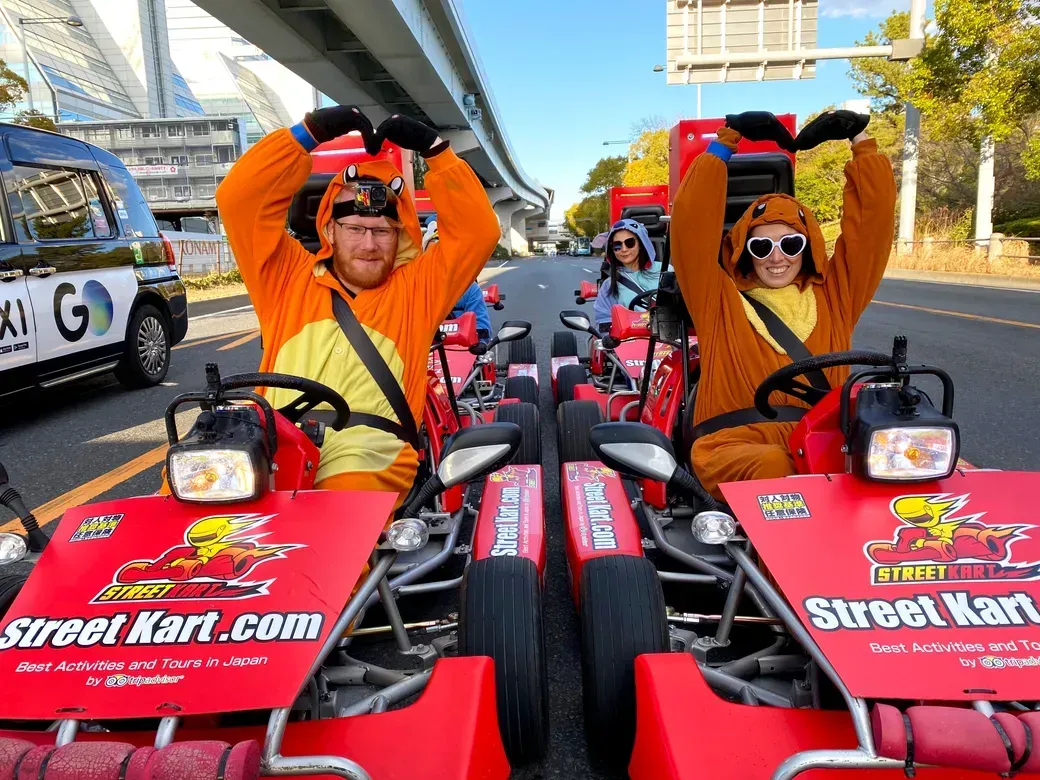 Two people in orange animal onesies pose in red go-karts on a city street, forming a heart shape with their arms; Street Kart branding on the front panels of the karts.