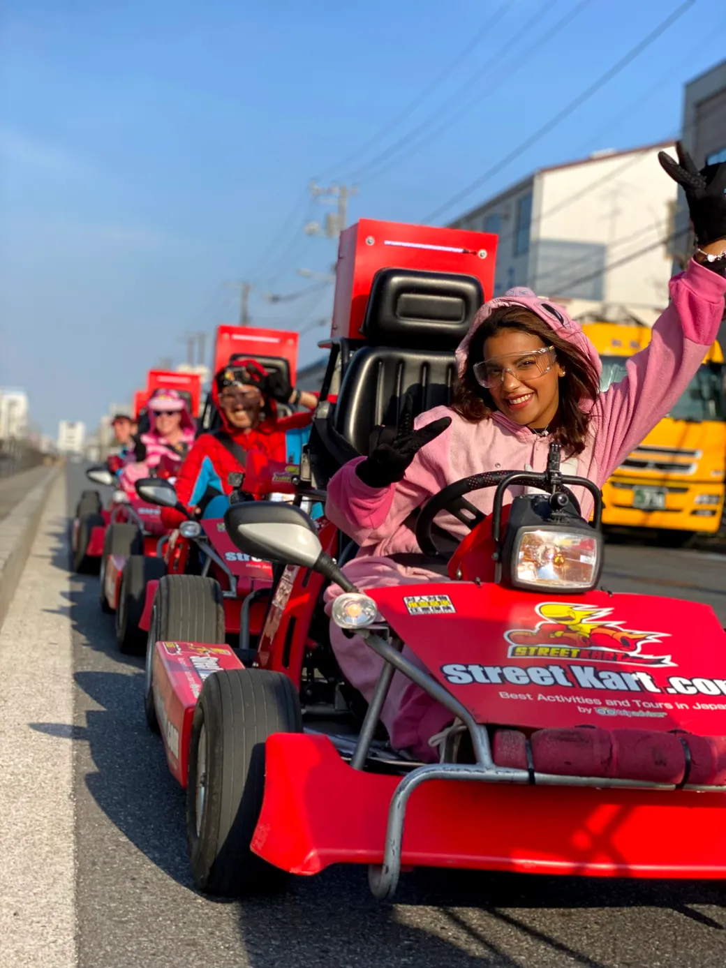Group of people in pink outfits riding red go-karts on a road, front rider waving and smiling with a Street Kart banner visible on the kart.